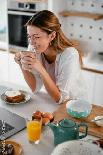 Young woman drinking coffee in kitchen. Beautiful woman having video call.
