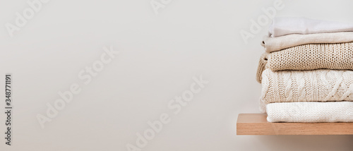 A stack of neatly folded warm knitwear, wool on a wooden shelf and a beige background. Capsule wardrobe, clothes storage, minimalism, knitted texture, order, comfort. Banner with copy space for text.