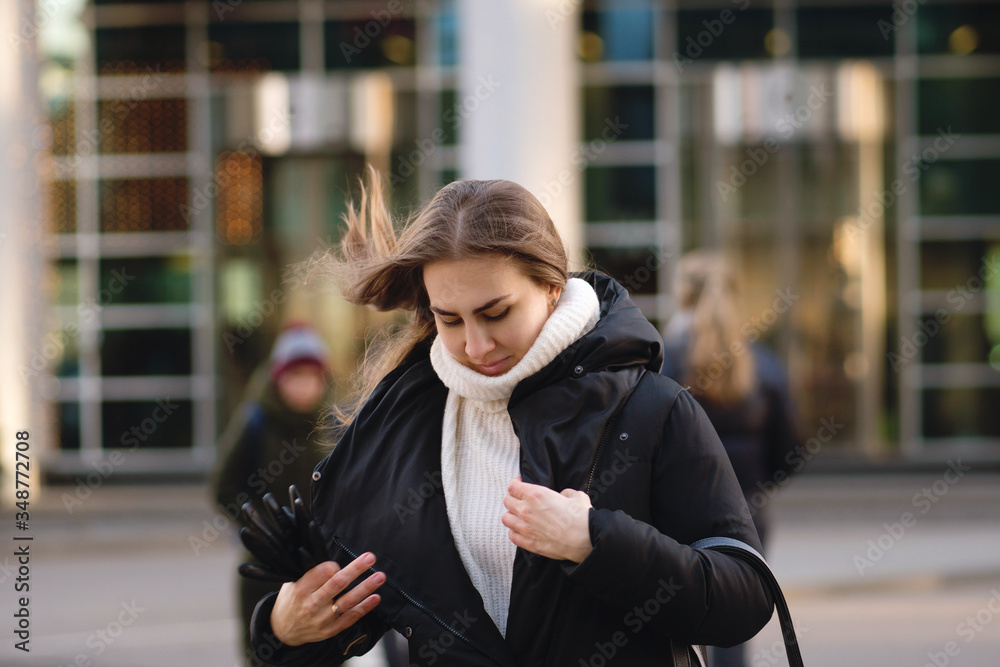 Fototapeta premium Portrait of a confident busy modern pretty woman. Girl hurries and fastens outerwear on the go. Hair develops in the wind. Background business center