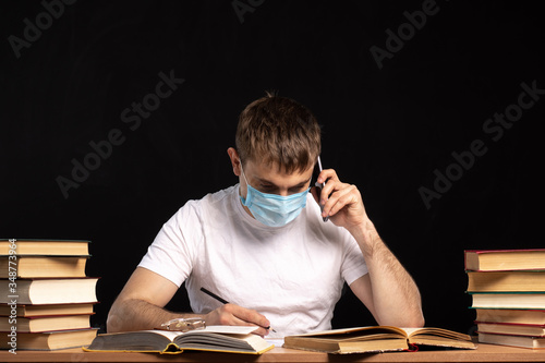 a young man in a medical mask is sitting at a table with books and talking on the phone. the difficulties of remote quarantined work. online training. Black background. infection protection