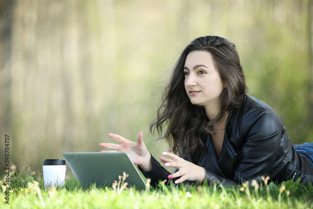 Obraz premium Young woman in a black jacked with coffee cup and laptop working outside in a park. Remote work. Digital work. Distance learning