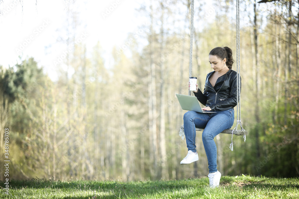 Young woman in a black jacked with coffee cup and laptop working outside in a park. Remote work. Digital work. Distance learning Young woman in a black jacked with coffee cup and laptop working outside in a park. Remote work. Digital work. Distance learning