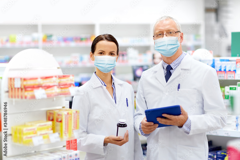 medicine, healthcare and technology concept - apothecaries wearing face protective medical mask for protection from virus disease with tablet pc computer and medication at pharmacy