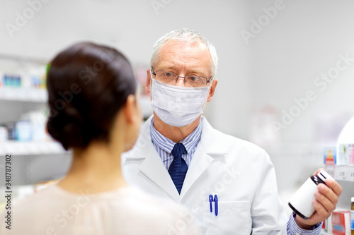 medicine, healthcare and people concept - senior apothecary wearing face protective medical mask for protection from virus disease showing drug to female customer at pharmacy