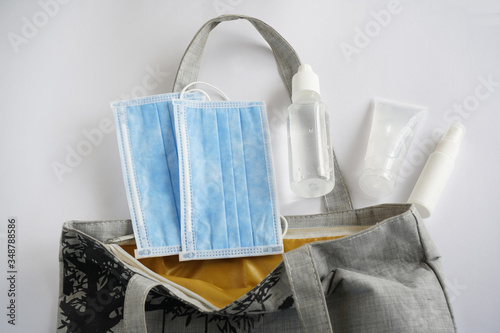 Blue face masks, a saline bottle, an alcohol gel and a spray are in an open textile bag. The items are essential to prevent Corona virus (Covid-19) during pandemic in 2020.    
