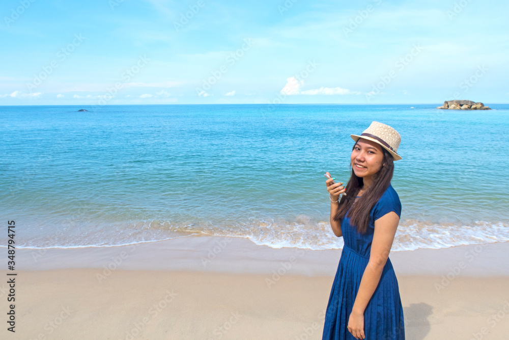 Cute Asian young teenager girl wearing sun hat & navy blue dress using mobile phone or smartphone taking photo or picture of deep blue sea with soft calm waves touching tropical summer sand beach Cute Asian young teenager girl wearing sun hat & navy blue dress using mobile phone or smartphone taking photo or picture of deep blue sea with soft calm waves touching tropical summer sand beach