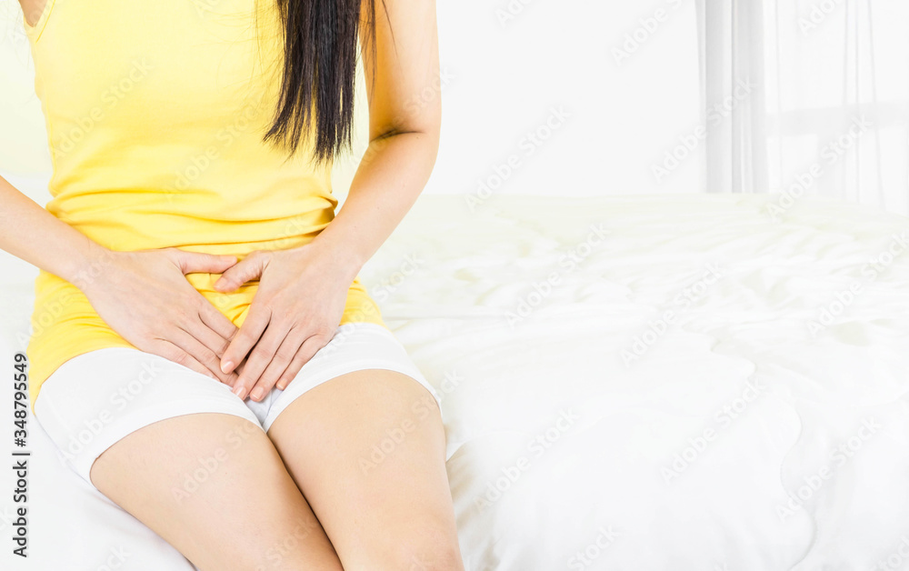 Obraz premium young asian woman wearing yellow undershirt Sitting on a white bed by the window with a thin curtain Female holding hand to crotch, Bladder pain,vagina-ache genital health care concept