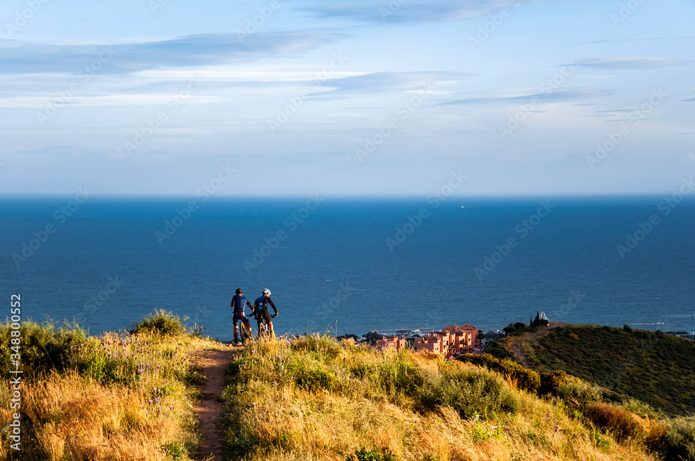 Two cyclists on a mountain road from the top of the hill look at the sea and the village below on the coast