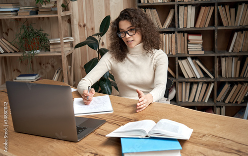 Virtual teaching concept. Female young hispanic school math teacher, college tutor coach looking at webcam and talking in classroom giving remote class online lesson by zoom conference call on laptop. photo