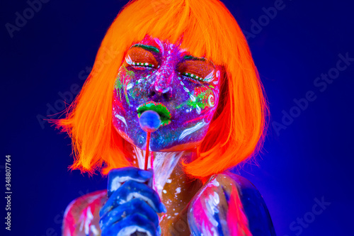 Fototapeta Naklejka Na Ścianę i Meble -  Fashion model woman licks sweet candy in neon light, disco night club. Beautiful model girl colorful bright fluorescent makeup