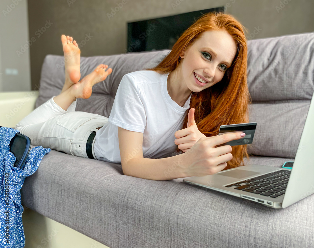 red-haired young woman is lying on a sofa with her legs up, shopping online on a laptop with a payment card in her hand. red-haired young woman is lying on a sofa with her legs up, shopping online on a laptop with a payment card in her hand.