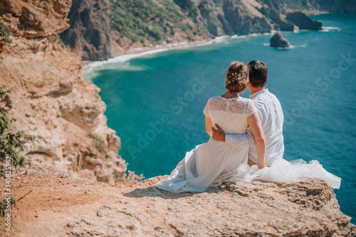 A young couple on vacation sitting with their arms around each other and looking at the beautiful seascape. In white wedding clothes. The concept of romance, travel, adventure.