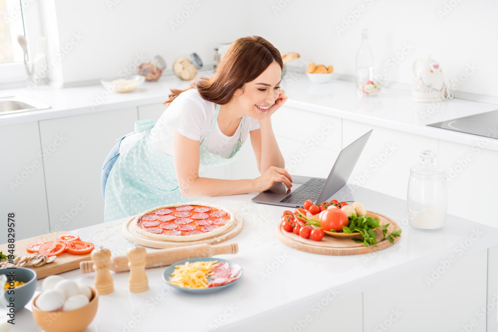 Portrait of charming cheerful girl want cook tasty pizza use laptop watch online video chef courses seminar wear dotted apron in house kitchen Portrait of charming cheerful girl want cook tasty pizza use laptop watch online video chef courses seminar wear dotted apron in house kitchen