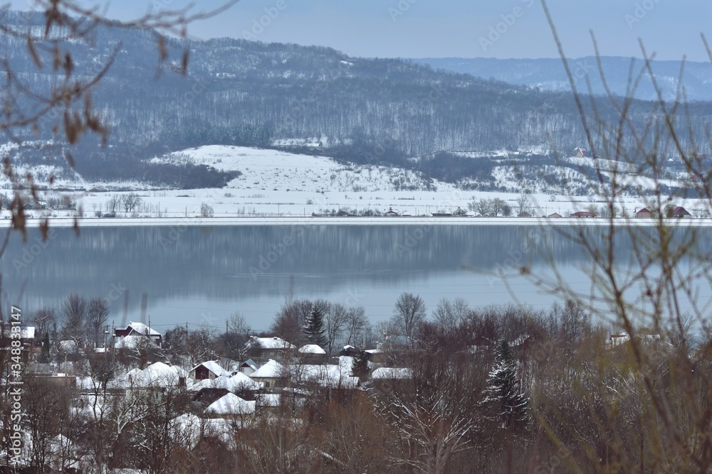 frozen lake in winter season close to the village with forest reflecting in water