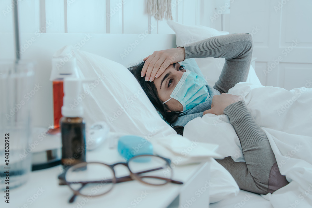 Young, sick woman lies tired in bed with a face mask on and holds her head. Young, sick woman lies tired in bed with a face mask on and holds her head.