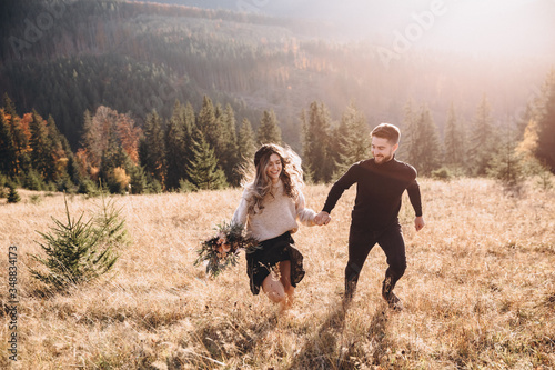 Stylish model couple in the autumn mountains. A young guy and a girl run along the slope against the background of the forest and mountain peaks at sunset. Girl holds in her hands a bouquet of flowers © AlexGukalovUkraine