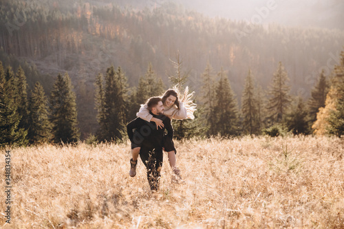 Stylish model couple in the autumn mountains. A young guy and a girl run along the slope against the background of the forest and mountain peaks at sunset.