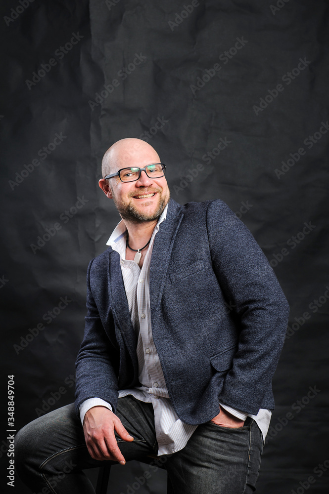 A young man in glasses and a jacket is smiling, looking at the camera and sitting on a bar stool against a black background. A young man in glasses and a jacket is smiling, looking at the camera and sitting on a bar stool against a black background.