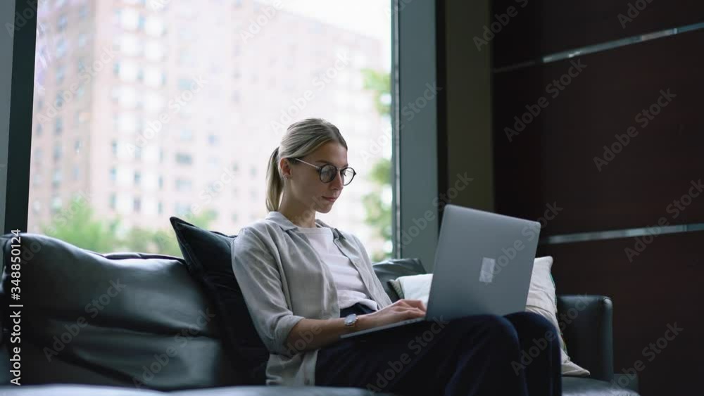 Young woman keyboarding information on website and publicating content in social networks on modern laptop using wireless internet connection while checking paper reports sitting in modern apartment
