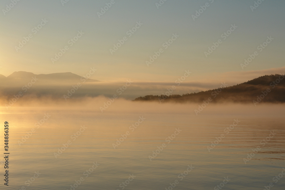 A scenic landscape - morning fog over the calm water of Chivyrkuisky Bay. Golden sunrise in a Zmeyevaya Bay, peninsula Svyatoy Nos (Holy Nose peninsula), Baikal Lake, Siberia, Russia