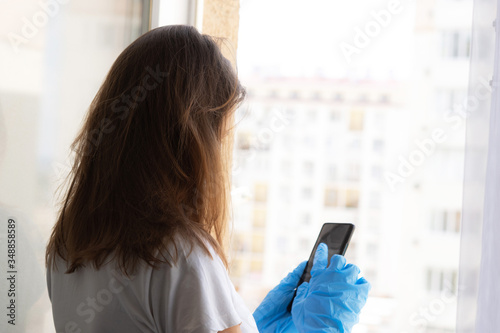 The girl at home at the window with the phone in her hands. A girl during quarantine due to the coronavirus pandemic looks out through a window. Isolation and self-isolation.