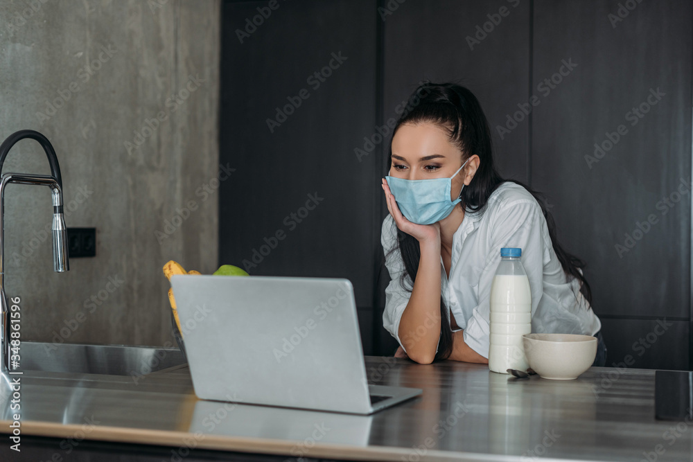 young woman in protective mask looking at laptop near bottle of milk in kitchen young woman in protective mask looking at laptop near bottle of milk in kitchen