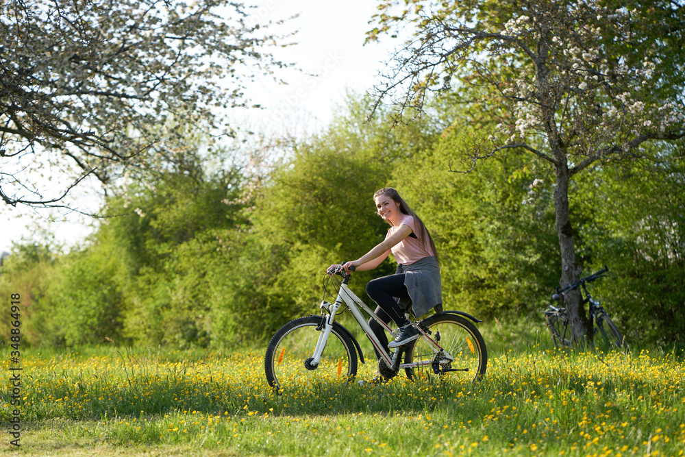 Smiling young woman on bicycle in a meadow with yellow flowers, in the background bushes and bicycles leaning on an apple tree
