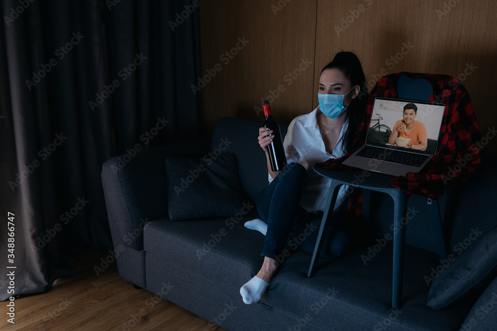 young woman in medical mask holding bottle of wine while sitting on sofa near laptop with smiling asian boyfriend on screen young woman in medical mask holding bottle of wine while sitting on sofa near laptop with smiling asian boyfriend on screen