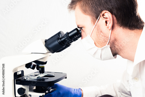 White male scientist using binocular microscope, wearing face mask and gloves.