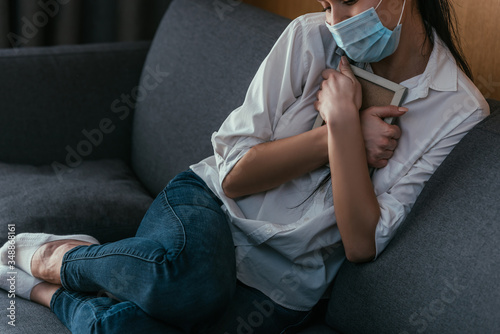 cropped view of depressed young woman in medical mask grieving while holding photo frame near chest while sitting on sofa