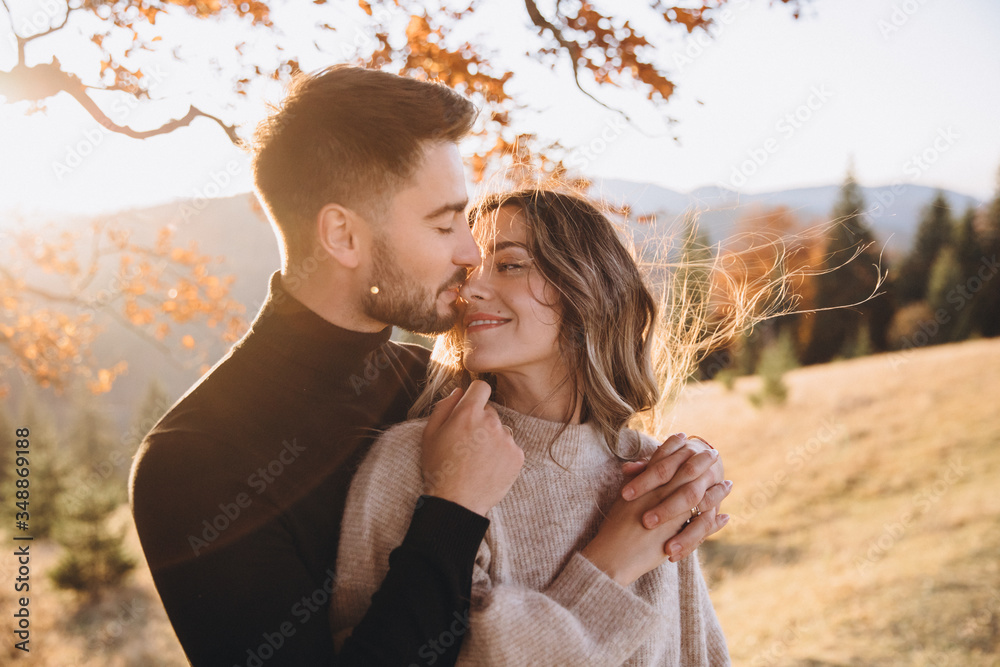 Fototapeta premium Stylish young couple in the autumn mountains. A guy and a girl hug together under a large old tree on a background of a forest and mountain peaks at sunset.