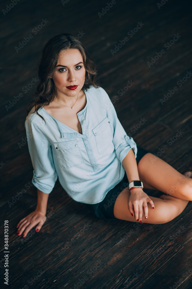 Φωτογραφία Beautiful young woman with perfect dark curly hair and beautiful make-up with chubby red lips in a denim shirt and shorts in a studio on a wooden floor