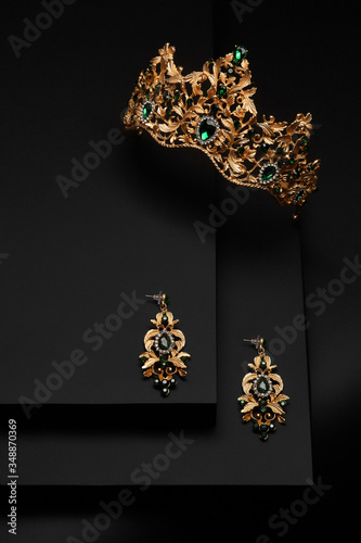 Subject shot of a golden tiara and chandelier earrings on the black stepped surface. The luxury jewelry set is adorned with emerald gems and rhinestones.