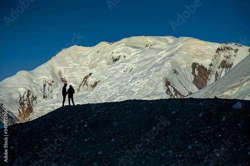 Scenic minimalistic landscape of snowy mountain peak. Blue sky. Silhouette of two men on the foreground.