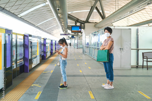 Asian woman wearing face mask and using phone and keep distance to other people while waiting subway in empty subway platform during coronavirus outbreak. social distancing, new normal concepts