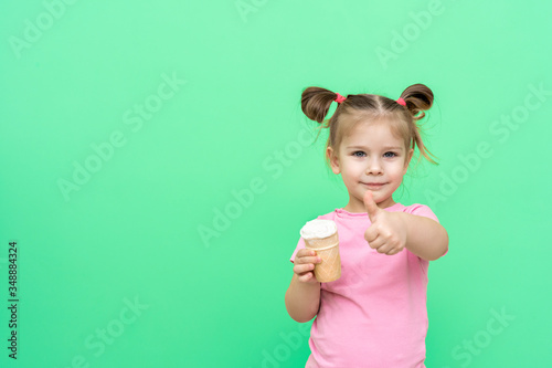little girl 4 years old in a pink T-shirt on a green background holds ice cream in his hand, with his other hand shows the sign OK, thumb up. layout with place for text