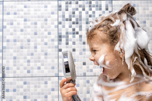 a little girl in foam is standing in the bathtub in her hand holding a shower and is yelling emotionally at him, there is no water. the child can not wash off the soap, turning off the water