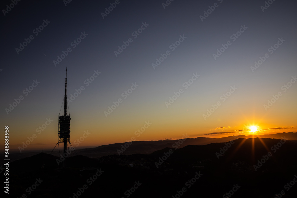 Big telecommunications antenna silhouette on the hills during sunset with sun rays