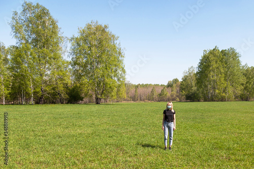 Fototapeta Naklejka Na Ścianę i Meble -  Girl blond in black T-shirt and blue jeans with a medical mask on her face is alone in the field against a background of trees and grass. Self-isolation regime in the context of a coronavirus pandemic