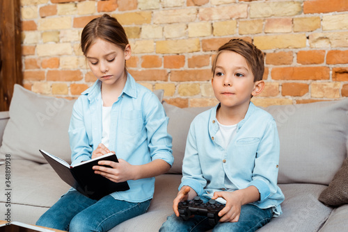 KYIV, UKRAINE - APRIL 27, 2020: sister writing in notebook near brother playing video game