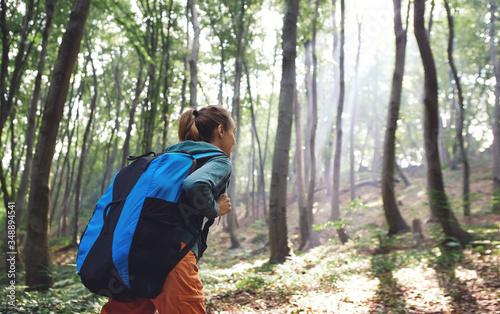 back view woman traveler with big backpack hiking walking on rocky trail throught forest, enjoying scenery of nature, sun rays shine through trees. Outdoor adventure, people in nature, solo traveling.