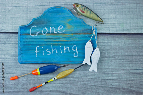 Gone fishing sign written on a pretty wooden plaque. Fishing lures and floats on rustic homemade woody background.Summer fishing flat lay. Top view with copy space