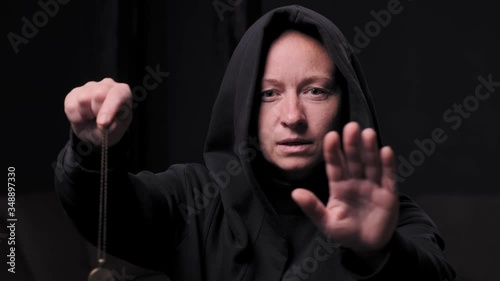witch woman swings pendant on chain while holding in hand in black room.