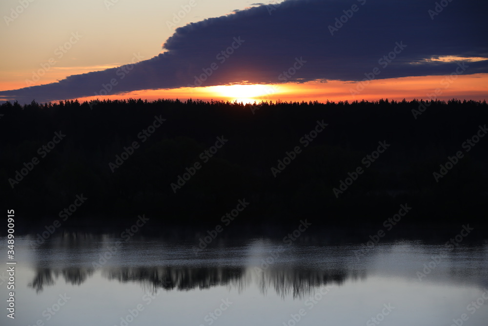 Dramatic sky background.The Golden light of the rising sun at dusk illuminates the overhanging continuous cloud on the clear horizon and the forest reflected in the ripples of a calm lake.Russia