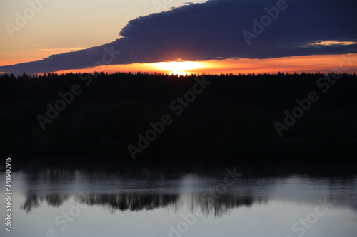 Dramatic sky background.The Golden light of the rising sun at dusk illuminates the overhanging continuous cloud on the clear horizon and the forest reflected in the ripples of a calm lake.Russia