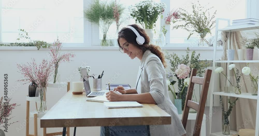 Female florist designer wears headphone elearning online watching video training course on laptop at work surrounded with flowers. Wedding planner video conferencing client in modern floristry studio.