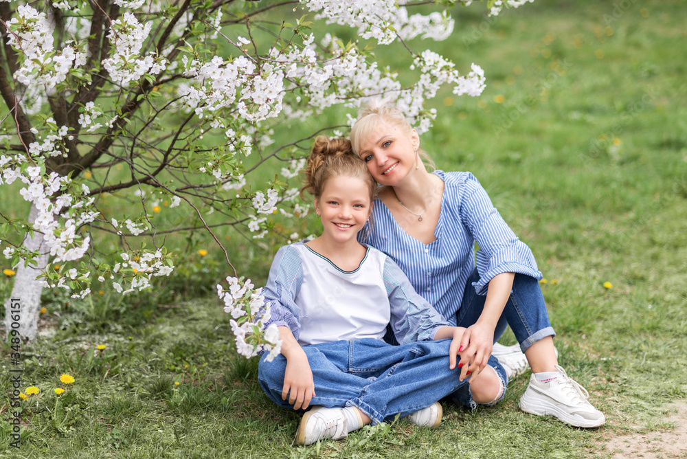 Fototapeta premium Happy mother and daughter sit on the grass in a blooming garden in spring
