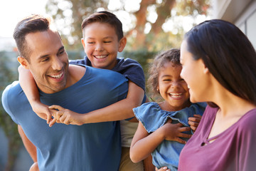 Smiling Hispanic Family With Parents Giving Children Piggyback Rides In Garden At Home
