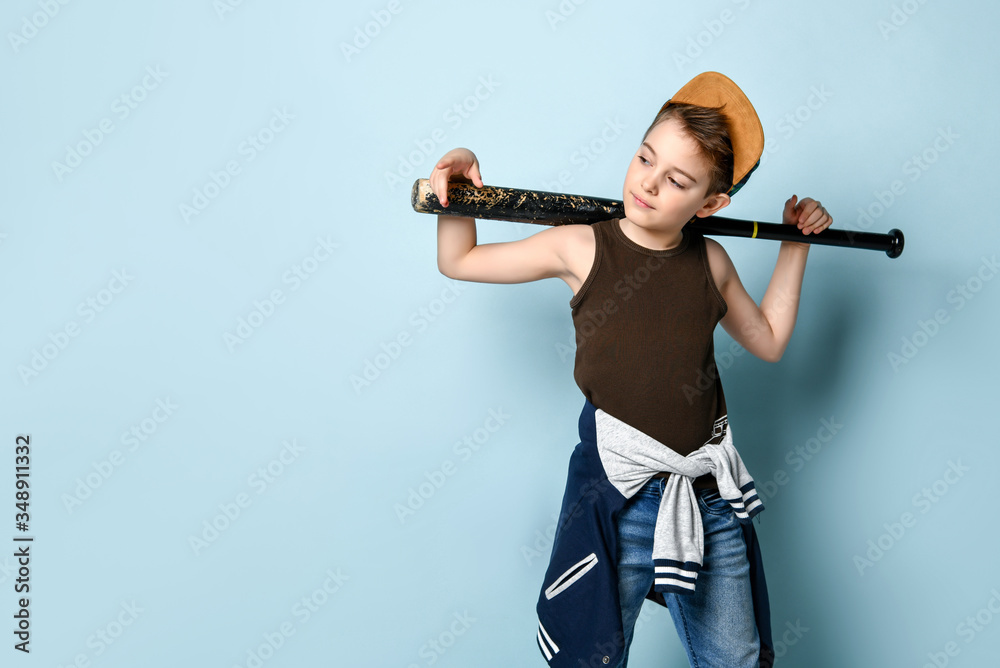 Sly little schoolboy stands near the wall looking aside holding baseball bat on shoulders. Three quarter length portrait isolated on blue background Sly little schoolboy stands near the wall looking aside holding baseball bat on shoulders. Three quarter length portrait isolated on blue background