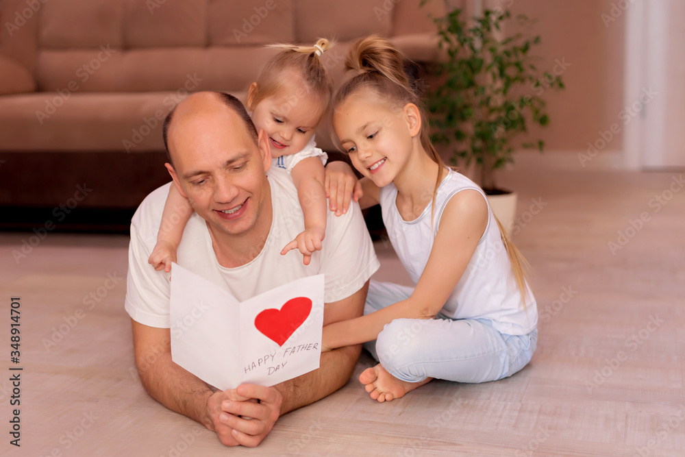 Happy young dad lying on the floor and holding a greeting card from little cheerful daughters on fathers day during home celebration Happy young dad lying on the floor and holding a greeting card from little cheerful daughters on fathers day during home celebration